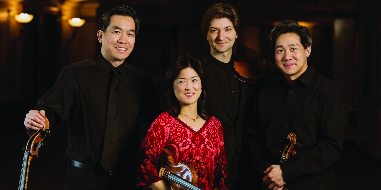Four musicians stand together, smiling, holding string instruments. Three men wear black shirts; one woman in a red patterned blouse stands in front. The background is dimly lit, giving a formal, concert-like atmosphere.