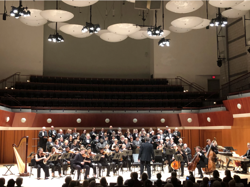 A symphony orchestra and choir perform on stage in a modern concert hall, with musicians playing strings and a conductor leading them. Audience members are visible in the foreground.