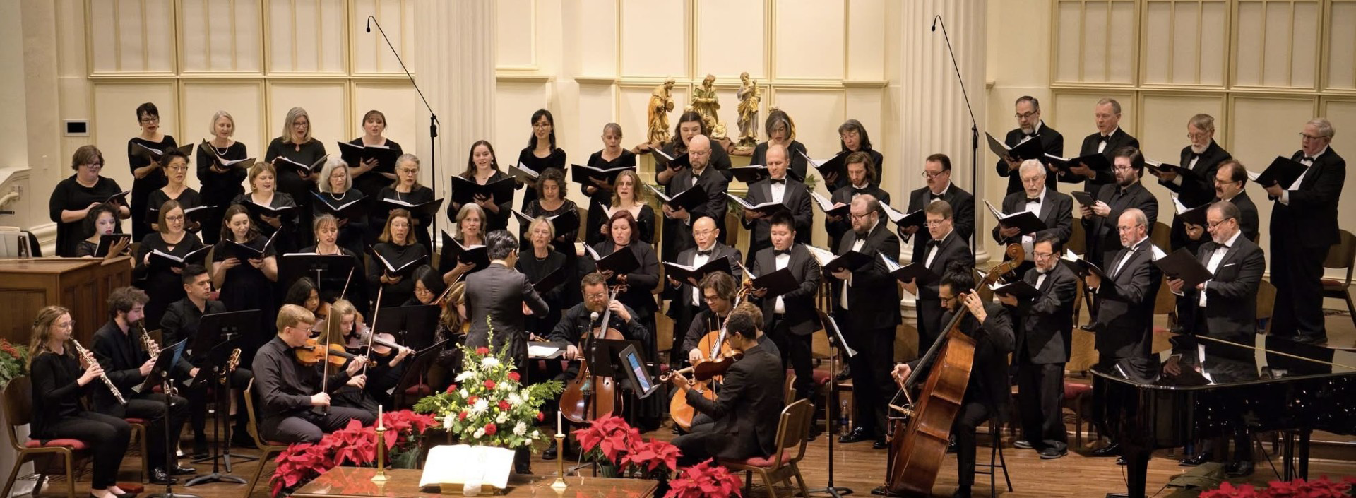 A choir and orchestra performing together on stage in a concert hall, with musicians playing various instruments and singers holding sheet music. The stage is decorated with flowers and red poinsettias.