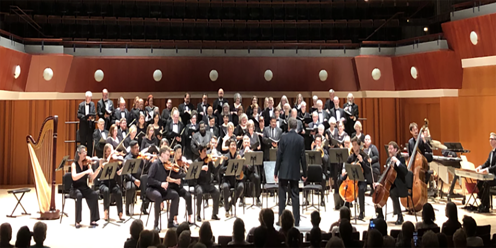 A symphony orchestra performs on stage with a conductor in front. Musicians play strings, harp, keyboard, and percussion. A large choir stands behind them, all in formal black attire, on a well-lit concert hall stage.