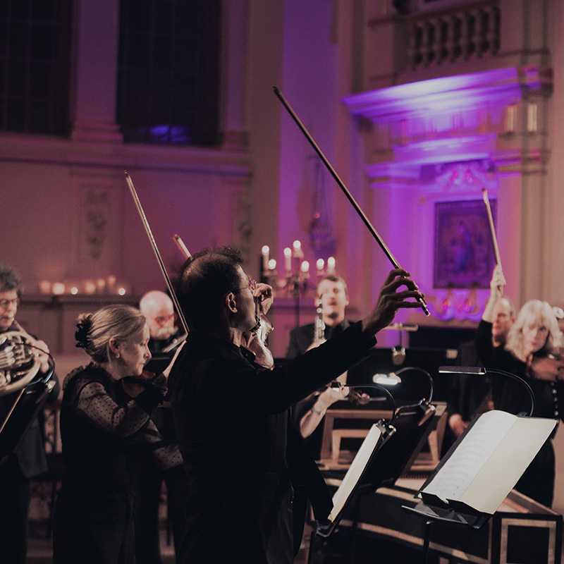 A group of classical musicians perform on stage in an elegant, candlelit hall; violinists and other instrumentalists are playing, with sheet music stands visible in the foreground.
