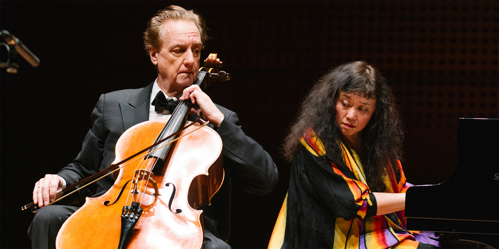 A man in a suit plays the cello while a woman in a colorful top plays the piano next to him on stage.