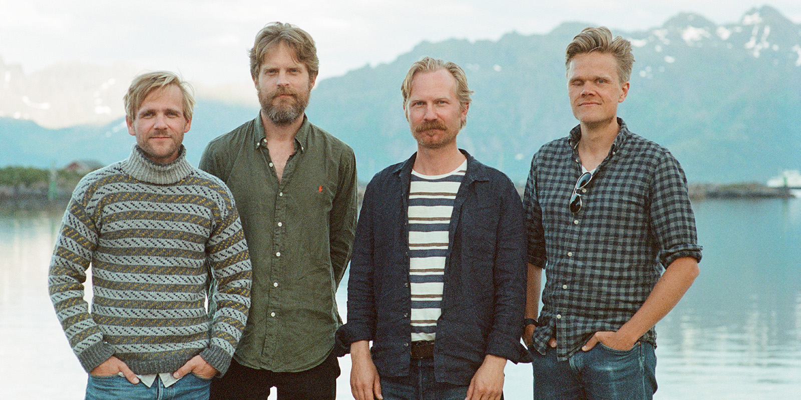 Four men stand side by side outdoors in front of a calm lake with mountains in the background. They wear casual shirts and sweaters, and all have light hair and beards. The weather appears mild and clear.