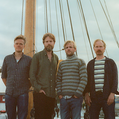 Four men stand side by side in front of a sailboat mast and rigging, with overcast skies in the background. They wear casual clothes, including shirts and sweaters, and look directly at the camera.