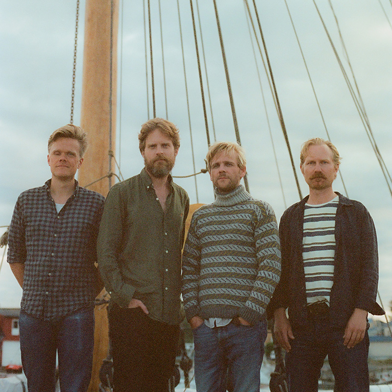 Four men stand together in front of a sailboat mast and rigging, posing outdoors. They wear casual clothing, including patterned shirts and sweaters, and have light hair and beards. The sky is cloudy in the background.