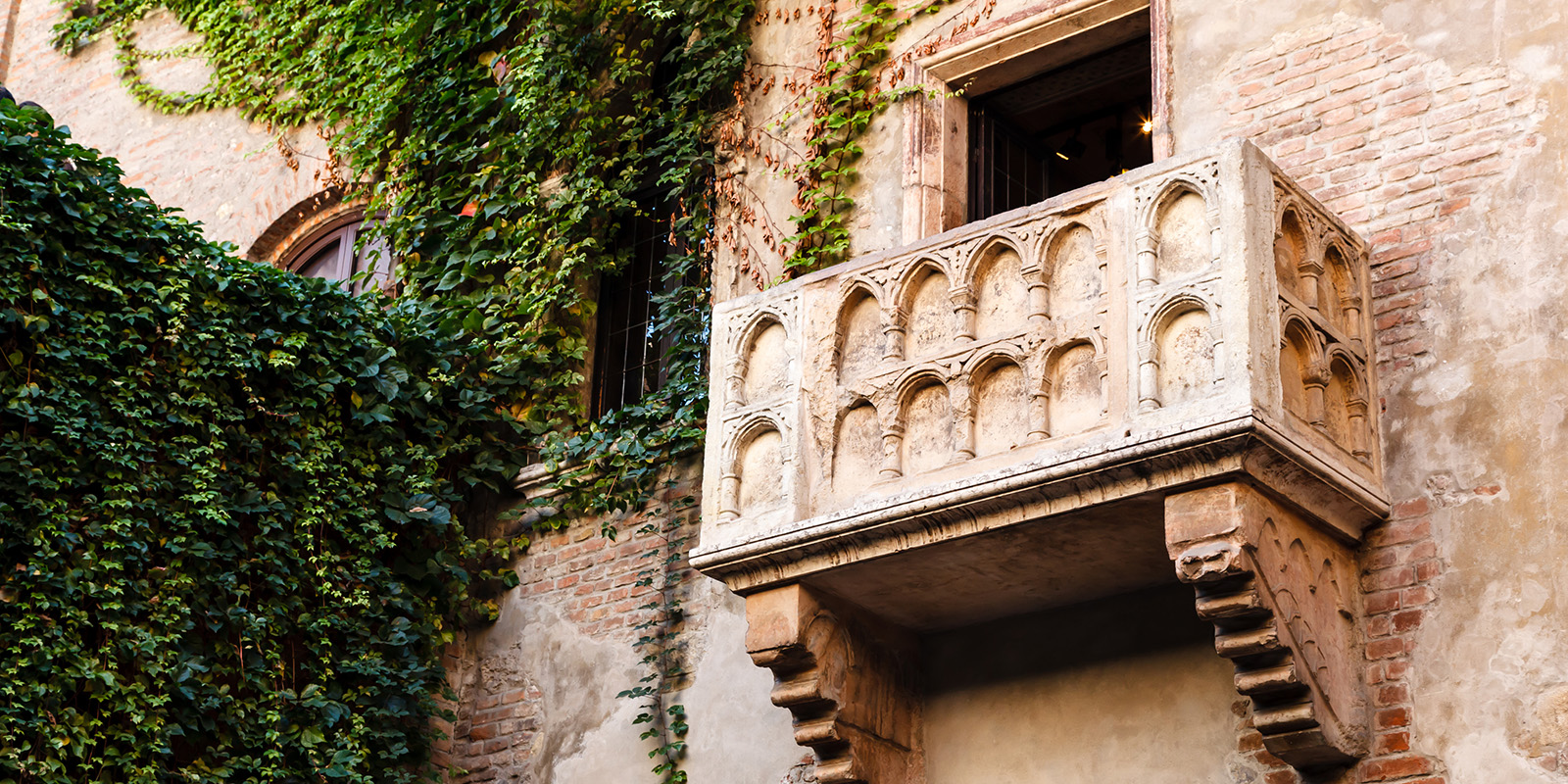 A stone balcony with detailed carvings juts from an old brick building covered in green ivy, next to an arched window.