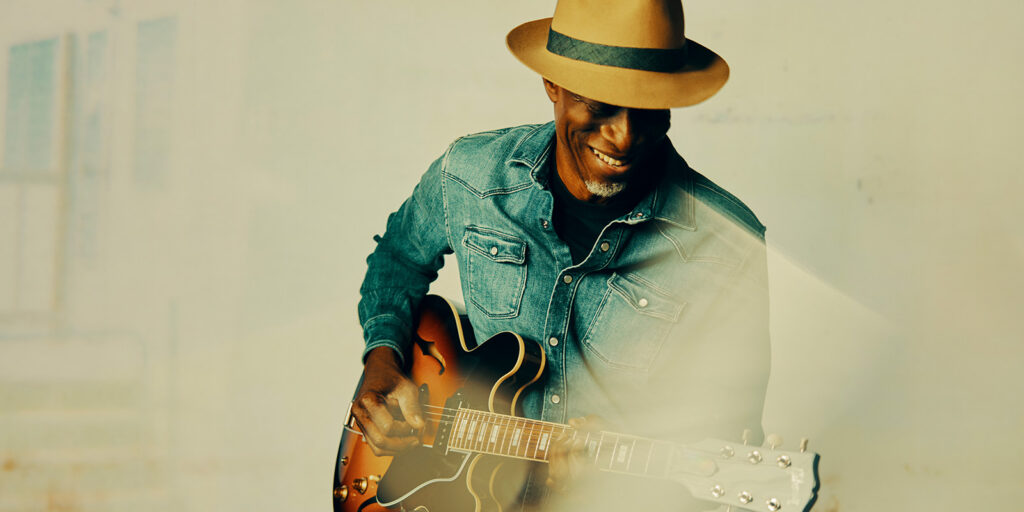 A smiling man in a tan fedora and denim shirt plays an electric guitar, standing against a light-colored background with a soft, dreamy effect.