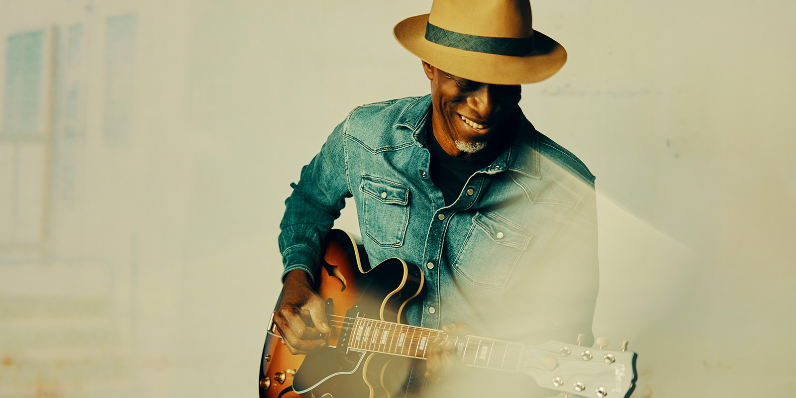A smiling man in a tan fedora and denim shirt plays an electric guitar, standing against a light-colored background with a soft, dreamy effect.