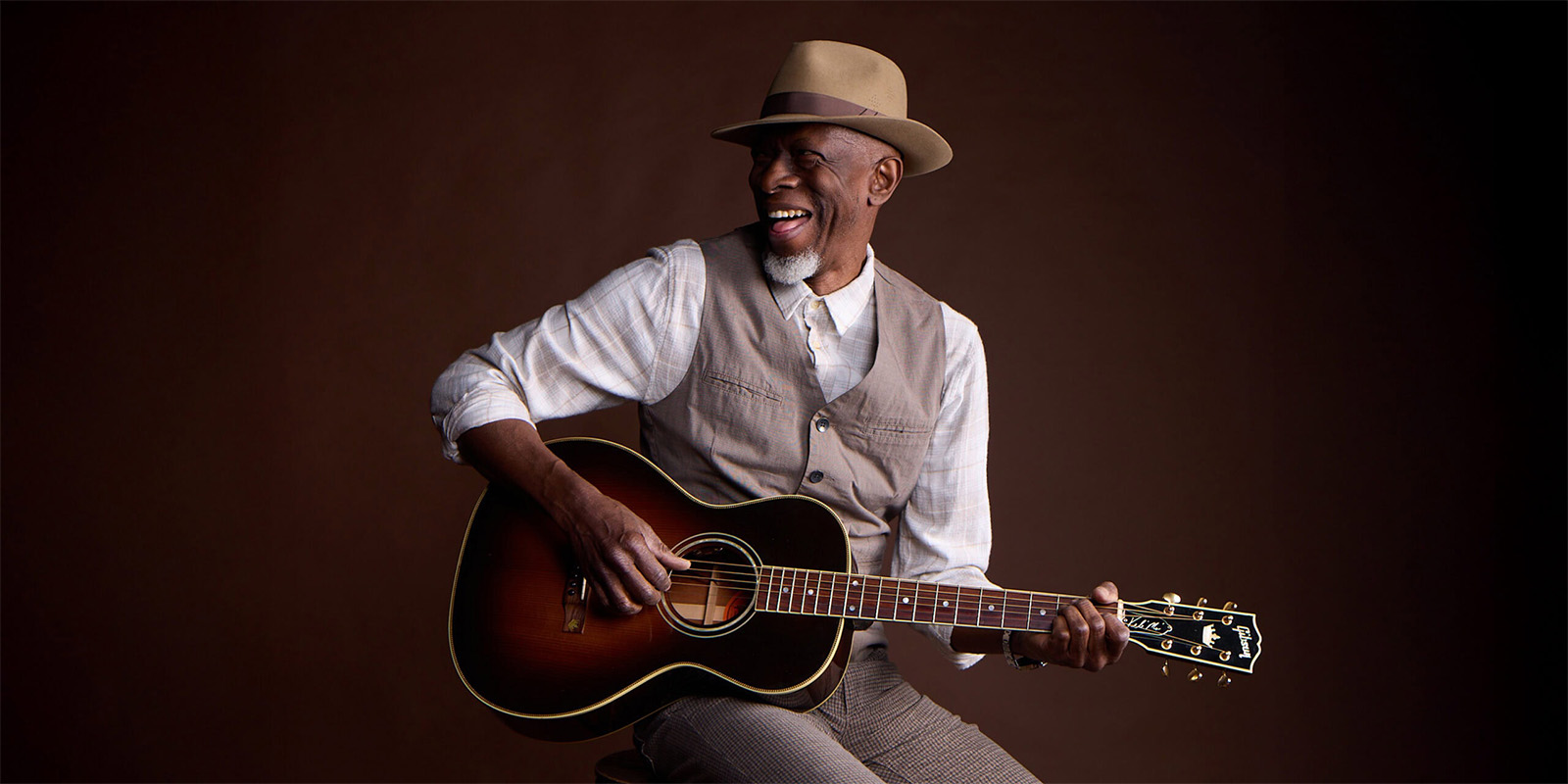 A smiling man in a beige hat and vest sits on a stool playing an acoustic guitar against a dark brown background.