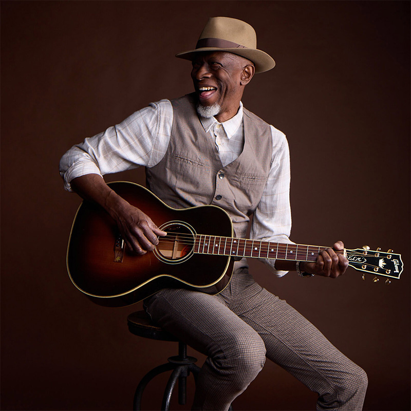 An older man in a hat, vest, and plaid pants sits on a stool, smiling while playing an acoustic guitar against a brown background.