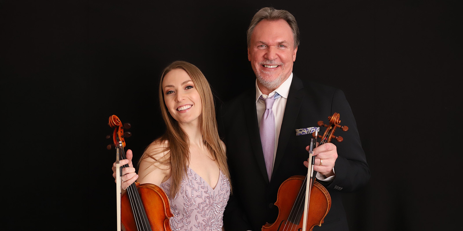 A smiling woman in a light purple dress and a man in a suit and tie stand side by side against a black background, each holding a violin.