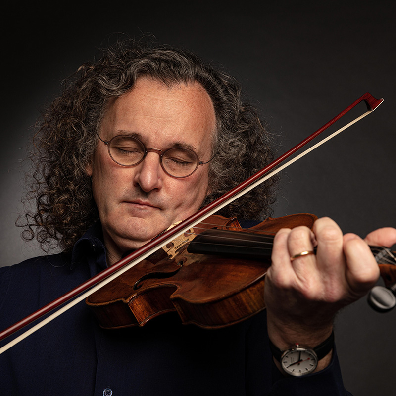 A man with curly hair and glasses plays a violin with his eyes closed, wearing a dark shirt and a watch, against a dark background.