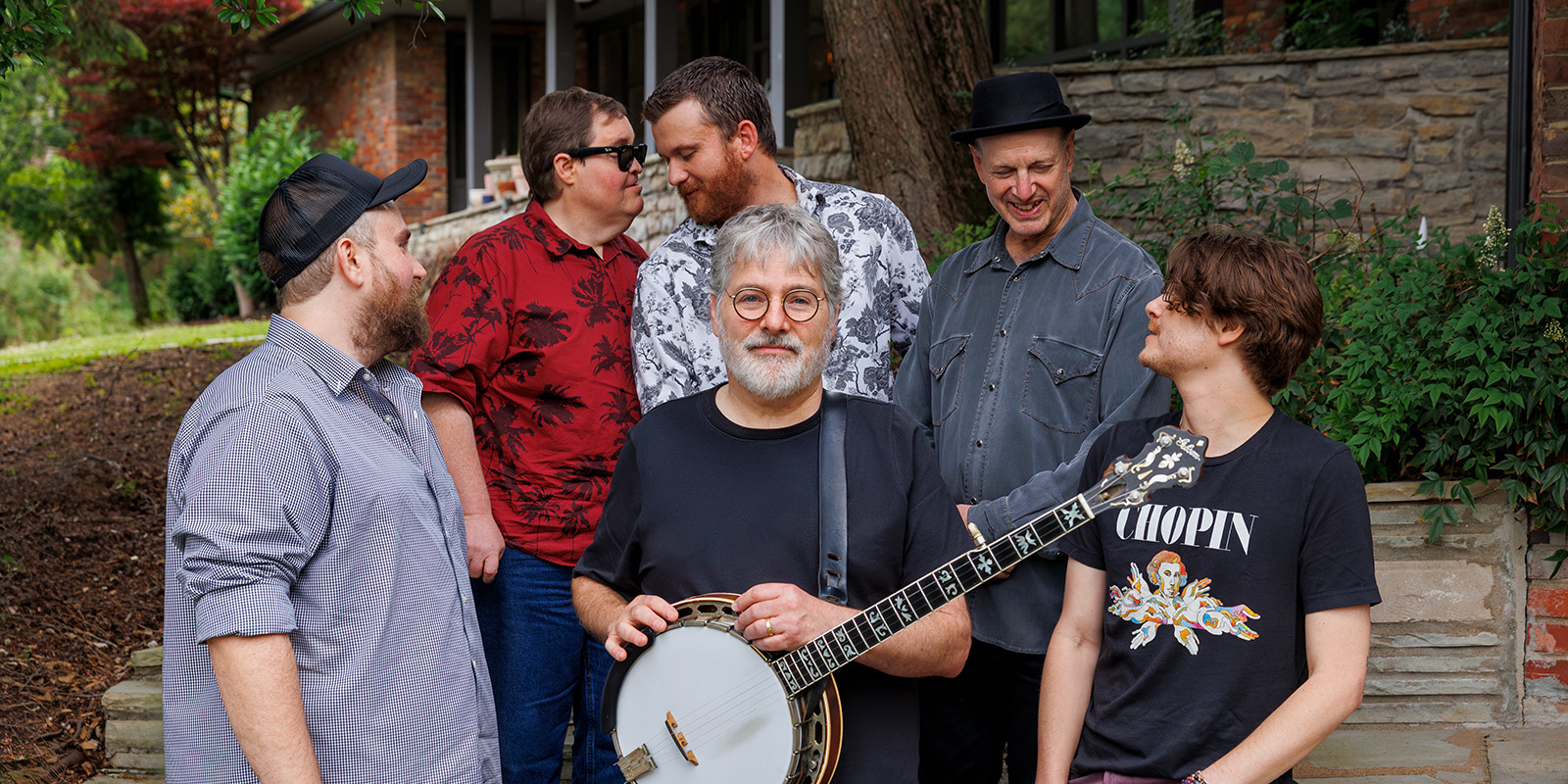 Six men are standing outdoors in front of a stone wall and greenery. One man in front holds a banjo and others stand casually around him, smiling and talking.