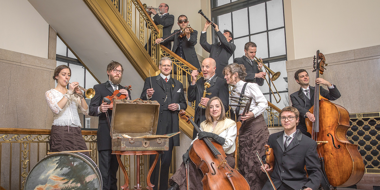A group of musicians in formal attire pose indoors with various instruments, including violin, trumpet, cello, drums, saxophone, clarinet, and double bass, standing on and around a staircase with gold railings.