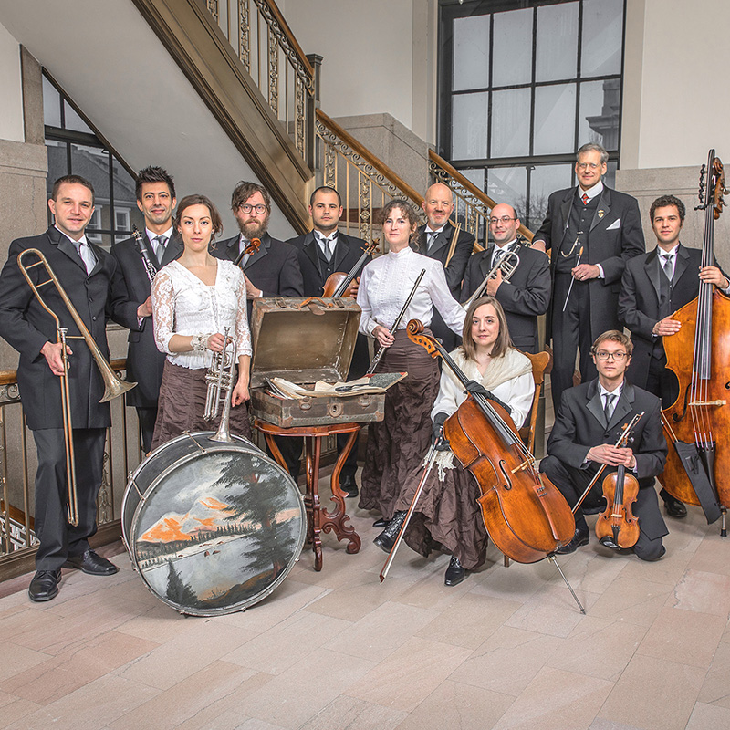 A group of musicians in formal attire pose with their instruments, including strings, brass, and percussion, in an elegant interior with a staircase and large windows in the background.