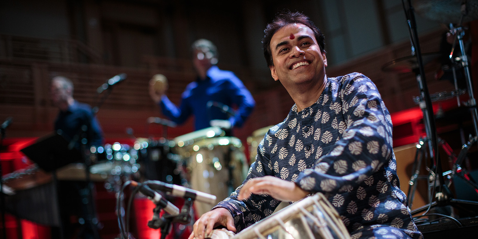 A smiling musician in patterned traditional attire plays a drum on stage, with two other musicians and percussion instruments in the background under colorful stage lighting.