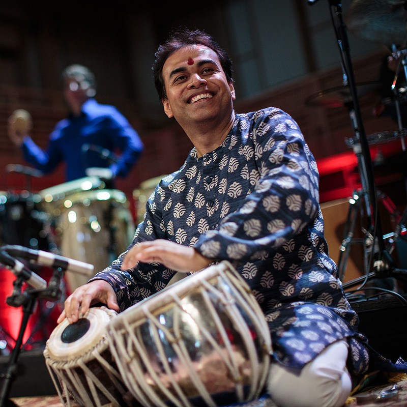 A smiling musician in traditional attire plays a tabla during a live performance, with another percussionist blurred in the background. The scene is set on a dimly lit stage with musical instruments visible.