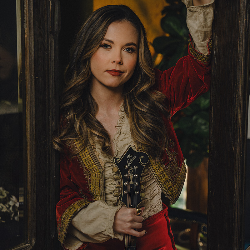 A woman with long wavy hair, wearing a red and gold embroidered jacket over a cream blouse, holds a string instrument and leans against a wooden door frame, looking at the camera.