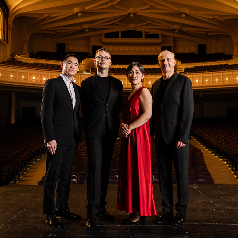 Four people stand on a theater stage, dressed formally. Three men wear black suits, and one woman wears a long red dress. The ornate, empty auditorium with warm lighting is visible behind them.