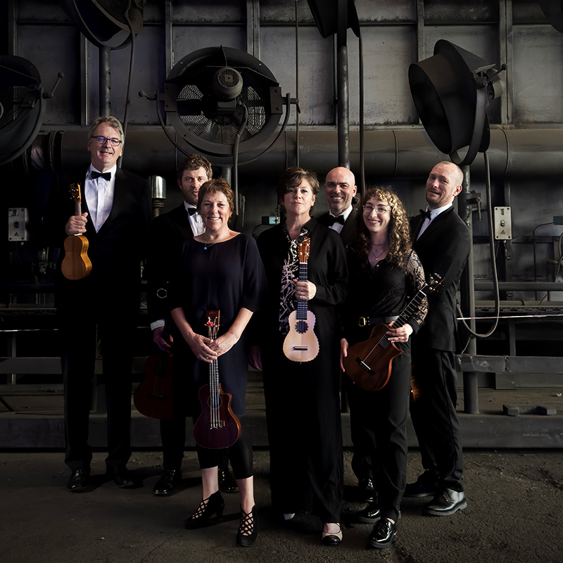 Seven people dressed in formal black attire stand in an industrial setting, each holding a string instrument, with large ventilation fans visible in the background. The group poses and smiles for the camera.