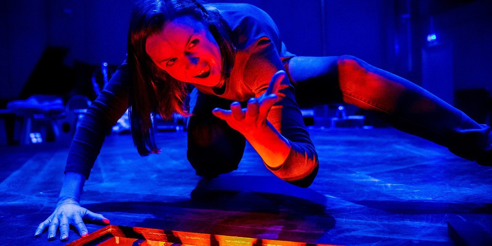 A person crouches on a dark stage, lit dramatically with blue and red lights, reaching forward with a fierce expression toward a glowing red opening in the floor.