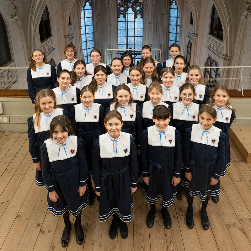 A group of girls in matching navy blue dresses with white collars and black shoes stand in rows inside a grand, historic building with arched windows and wooden floors.