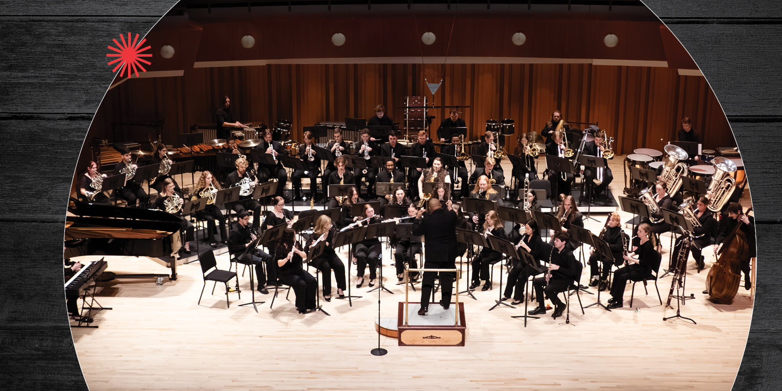 A large concert band with musicians playing various instruments is performing on stage in a concert hall, directed by a conductor at the center.