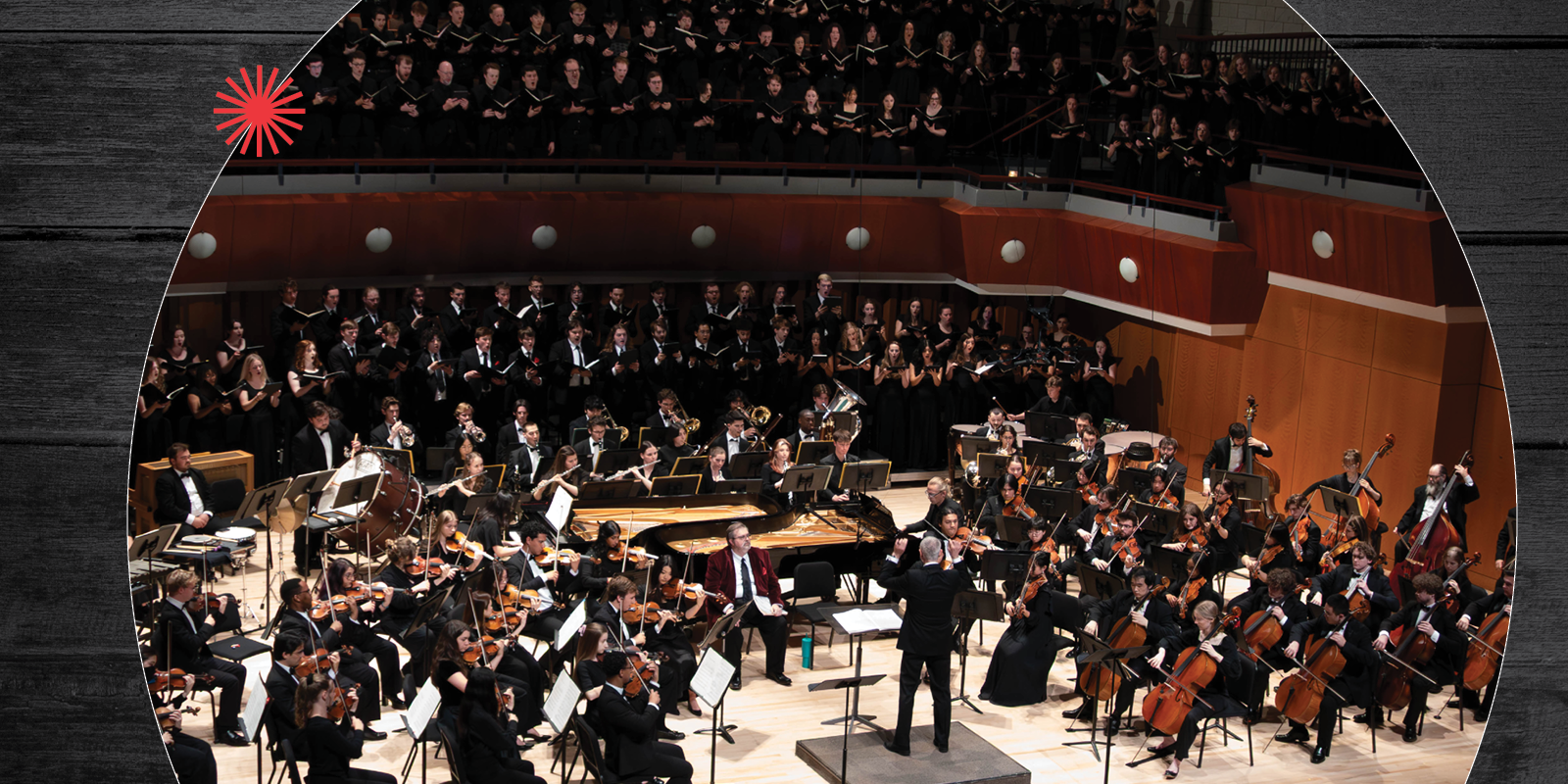 A large orchestra and choir perform on stage in a concert hall, with musicians seated in front playing various instruments and singers standing in rows behind them. A conductor stands at the center.