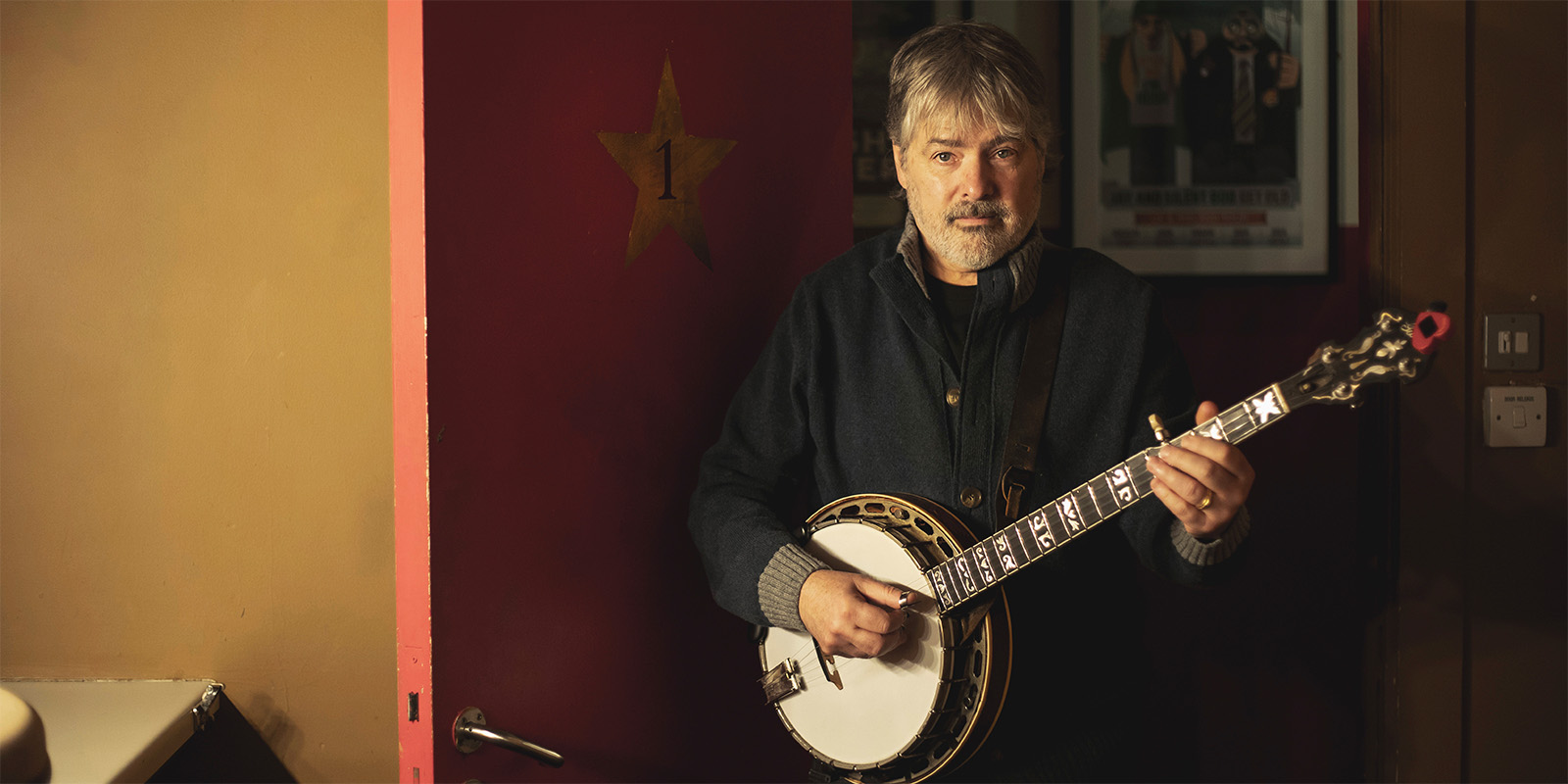 A man with gray hair and a beard stands indoors by a red door with a gold star, playing a banjo. He wears a dark jacket and looks toward the camera.