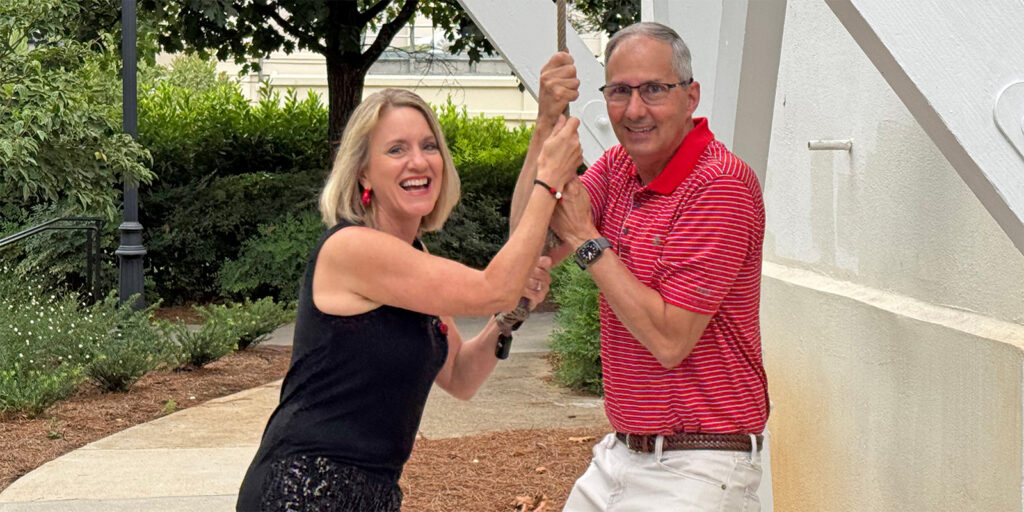 Two adults smiling and holding a large metal lever together outdoors, with greenery and a white structure in the background.